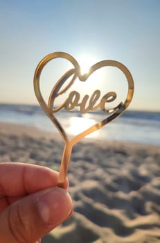 Person holding in fingers hand stick in shape golden heart and word Love on background sea and sea waves in seashore on sunny summer day close-up. Concept love romance amour St Valentines Day