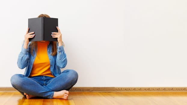 A young woman covers her face with a book as she sits on the floor against the wall.