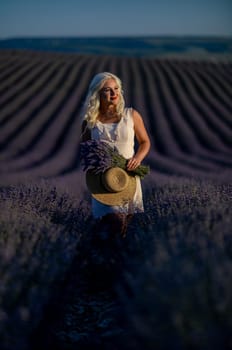 Blonde woman poses in lavender field at sunset. Happy woman in white dress holds lavender bouquet. Aromatherapy concept, lavender oil, photo session in lavender.