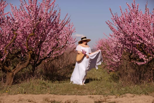 Woman blooming peach orchard. Against the backdrop of a picturesque peach orchard, a woman in a long white dress and hat enjoys a peaceful walk in the park, surrounded by the beauty of nature