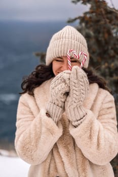 Woman candy sea. Smiling woman in knitted hat, mittens and beige coat holding lollipops candy canes in her hands in shape of heart against the backdrop of the sea