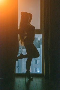 A full-length silhouette of young woman at open balcony window and dark blue night sky, with a cityscape in the background. Seduction and passion concept. Romantic moment in the bedroom. Night room