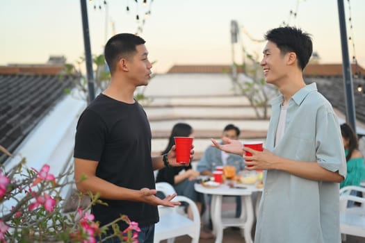 Two young handsome men having drinks and chatting at the rooftop party in the evening