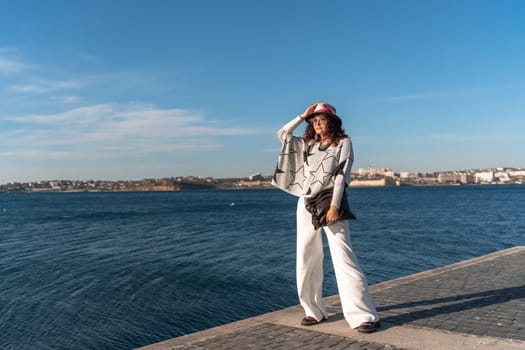 Stylish seashore woman. Fashionable woman in a hat, white trousers and a light sweater with a black pattern on the background of the sea