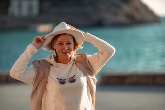 Happy blonde woman in a white suit and hat posing at the camera against the backdrop of the sea.