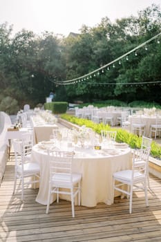 Festive tables with bouquets of flowers stand in the garden on a wooden flooring among the trees. High quality photo