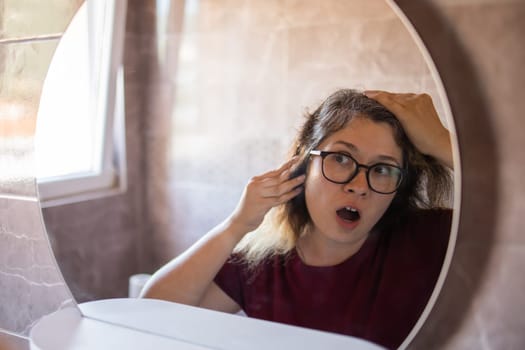 Woman Looking for Gray Hair in the Mirror. Young woman scared of premature aging signs.