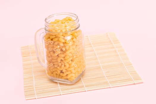 Uncooked Chifferi Rigati Pasta in Glass Jar on Bamboo Mat on Pink Background. Fat and Unhealthy Food. Scattered Classic Dry Macaroni. Italian Culture and Cuisine. Raw Pasta