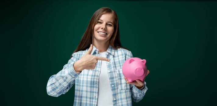 Young pretty woman holding pink piggy bank with savings on green background.