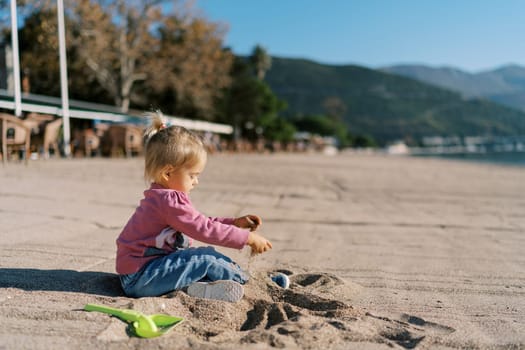 Little girl sits on the beach and pours sand with her hands. Side view. High quality photo