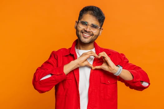 Man in love. Smiling attractive Indian man makes heart gesture demonstrates love sign expresses good positive feelings and sympathy. Handsome Arabian Hindu young guy isolated on orange wall background
