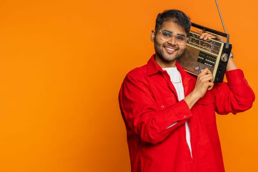 Happy young Indian man using retro tape record player to listen music, disco dancing favorite track, having fun entertaining, fan of vintage technologies. Arabian guy on orange background. Copy-space