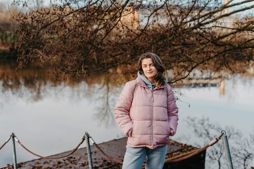 A young woman standing at the shore looking at the river in autumn sunny day. Street view, copy space for text, travel photo. Happy tourist woman on the bank of the river in autumn in warm clothes. Tourists enjoy their vacation, winter season. Romantic look and travel concept. A joyful mood in a Caucasian girl. Winter Wonderland: Enchanting Girl by the Riverside in Autumn.