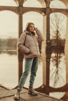 A young woman standing at the shore looking at the river in autumn sunny day. Street view, copy space for text, travel photo. Happy tourist woman on the bank of the river in autumn in warm clothes. Tourists enjoy their vacation, winter season. Romantic look and travel concept. A joyful mood in a Caucasian girl. Winter Wonderland: Enchanting Girl by the Riverside in Autumn.