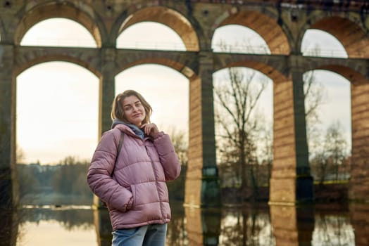 A young woman standing at the shore looking at the river in autumn sunny day. Street view, copy space for text, travel photo. Happy tourist woman on the bank of the river in autumn in warm clothes. Tourists enjoy their vacation, winter season. Romantic look and travel concept. A joyful mood in a Caucasian girl. Winter Wonderland: Enchanting Girl by the Riverside in Autumn.