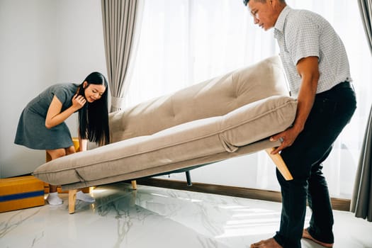 A portrait of a smiling Asian couple relocating joyfully moving a sofa in their new apartment. Ideal for depicting the excitement of furniture placement and home settling. Movinng day