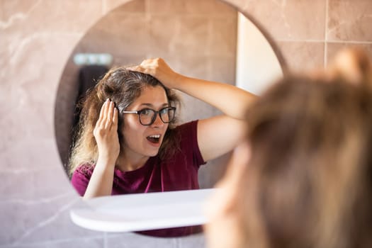 Gray haired surprised caucasian middle aged woman looking at grey hair head in mirror reflection