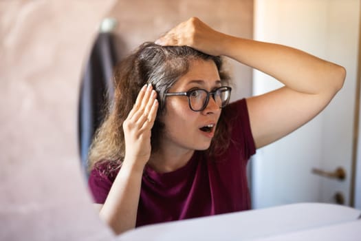 Gray haired surprised caucasian middle aged woman looking at grey hair head in mirror reflection