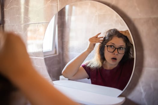 Gray haired surprised caucasian middle aged woman looking at grey hair head in mirror reflection