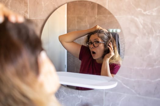 Gray haired surprised caucasian middle aged woman looking at grey hair head in mirror reflection