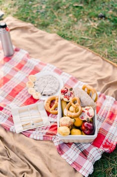 Basket of buns and fruit stands on a blanket next to cutlery and a thermos. High quality photo