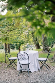 Table with a white tablecloth and a bouquet of flowers stands surrounded by chairs in the park. High quality photo