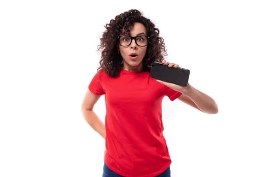 young active curly brunette woman dressed in a red basic t-shirt holding a smartphone.