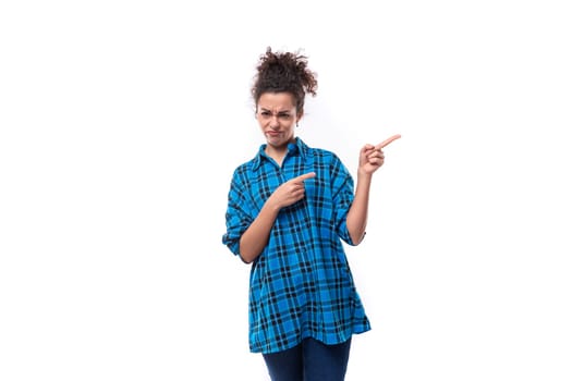 young dark haired european woman dressed informally in a blue plaid shirt points with her hand to the side isolated on white background.
