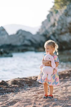 Little smiling girl stands on the beach, hugging a toy and looking at the sea. High quality photo