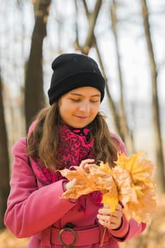 Portrait of beautiful happy young woman in pink jacket holding bouquet of yellow leaves at autumn park. Pretty Caucasian lady smiling and looking at camera during her walk outdoors. Generation Z and gen z youth.