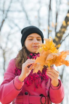 Portrait of beautiful happy young woman in pink jacket holding bouquet of yellow leaves at autumn park. Pretty Caucasian lady smiling and looking at camera during her walk outdoors. Generation Z and gen z youth.