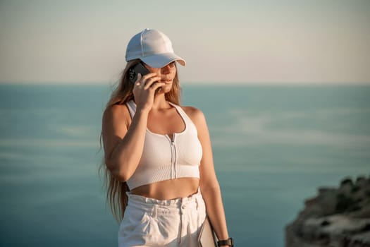 Freelance women sea working laptop. Happy middle aged woman talking on the phone outdoors with a beautiful view of the sea in her hand with a laptop. Remote work concept.