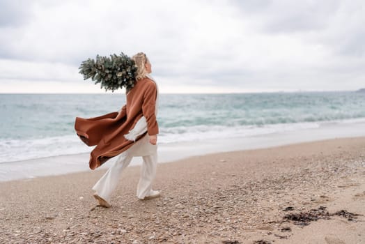 Blond woman Christmas sea. Christmas portrait of a happy woman walking along the beach and holding a Christmas tree on her shoulder. She is wearing a brown coat and a white suit