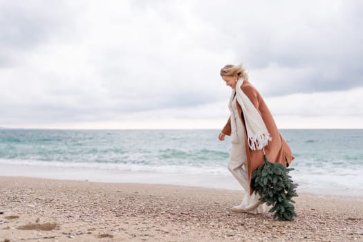 Blond woman Christmas sea. Christmas portrait of a happy woman walking along the beach and holding a Christmas tree in her hands. She is wearing a brown coat and a white suit