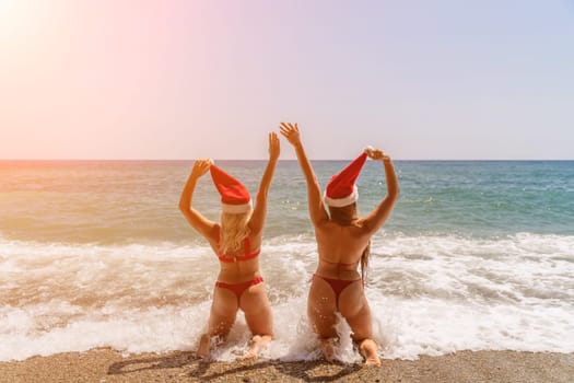 Women Santa hats ocean play. Seaside, beach daytime, enjoying beach fun. Two women in red swimsuits and Santa hats are enjoying themselves in the ocean, kneeling in the waves and raising their hands up