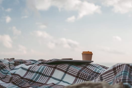 Laptop coffee on blanket with ocean view. Illustrating serene outdoor laptop use. Freelancer enjoying their time outdoors while working or browsing the internet