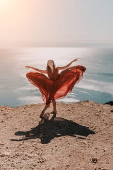 Woman red dress sea. Female dancer in a long red dress posing on a beach with rocks on sunny day. Girl on the nature on blue sky background