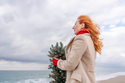 Redhead woman Christmas tree sea. Christmas portrait of a happy redhead woman walking along the beach and holding a Christmas tree in her hands. Dressed in a light coat, white suit and red mittens