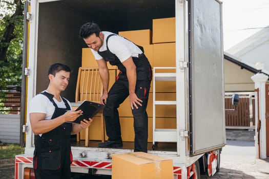 Mover workers in uniform unload boxes checking checklist with a clipboard at the truck. Professional delivery service ensures smooth relocation and efficient shipping. Moving day concept