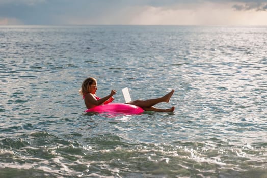 Woman works on laptop in sea. Freelancer, blond woman in sunglases floating on an inflatable big pink donut with a laptop in the sea at sunset. People summer vacation rest lifestyle concept