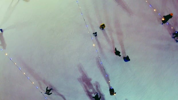 People skating on ice skating rink outdoors on winter night top view. Aerial drone view. Luminous glowing garlands lamps lighting. Cast long shadows. New Year Christmas celebration holidays recreation