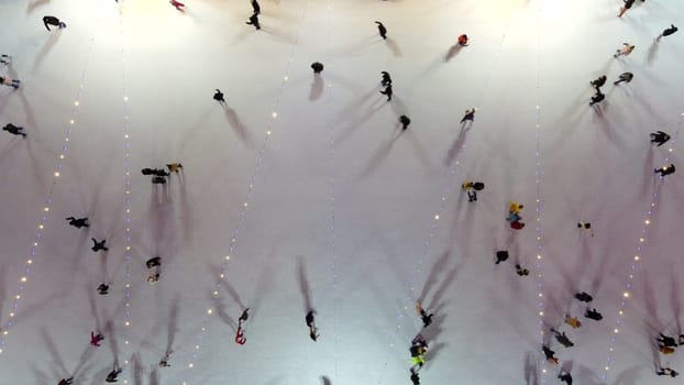 People skating on ice skating rink outdoors on winter night top view. Aerial drone view. Luminous glowing garlands lamps lighting. New Year Christmas celebration holidays recreation. Lowering down