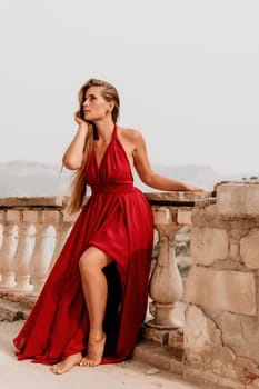 Side view a Young beautiful sensual woman in a red long dress posing on a volcanic rock high above the sea during sunset. Girl on the nature on overcast sky background. Fashion photo