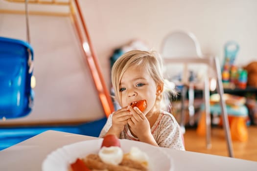 Little girl sits at the table and eats a piece of tomato. High quality photo
