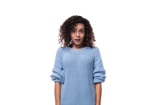 surprised emotional young curly brunette woman dressed in a blue sweater on a background with copy space.