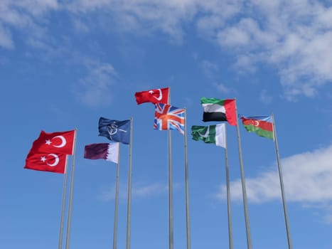 A striking arrangement of different national flags, such as Turkey, NATO, Qatar, the UK, the UAE, and Azerbaijan, presented in a row against a backdrop of a serene blue sky.
