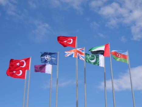 An array of flags from Turkey, Qatar, the United Kingdom, the United Arab Emirates, and Azerbaijan, along with the NATO emblem, captured in motion under the bright sunlight.