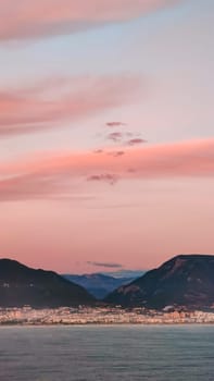 Pink clouds in evening sky over blue sea on background of city and mountains on horizon, vertical frame.