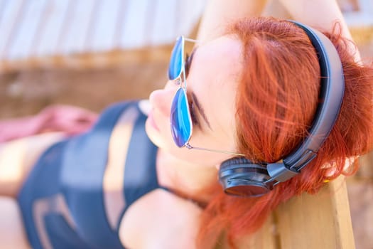 Cute woman listening to music on the beach. Red-haired young woman lying on the beach, relaxing on a lounger and listening to relaxing music.