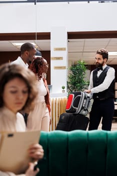 Male employee helping clients with bags, carrying luggage for people after doing check in procedure. Young couple receiving assistance from luxury resort bellboy at reception desk.